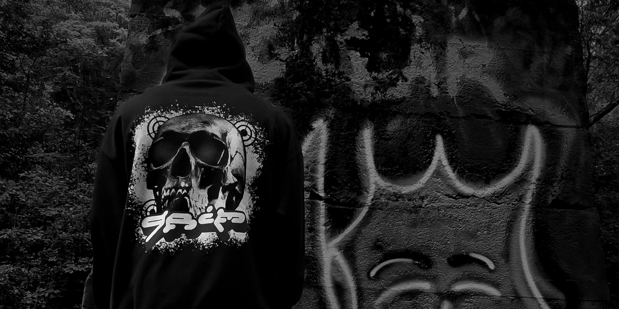 Black streetwear hoodie with large skull design by KIBISHI, styled in front of urban graffiti wall — artistic and edgy clothing for modern street culture.